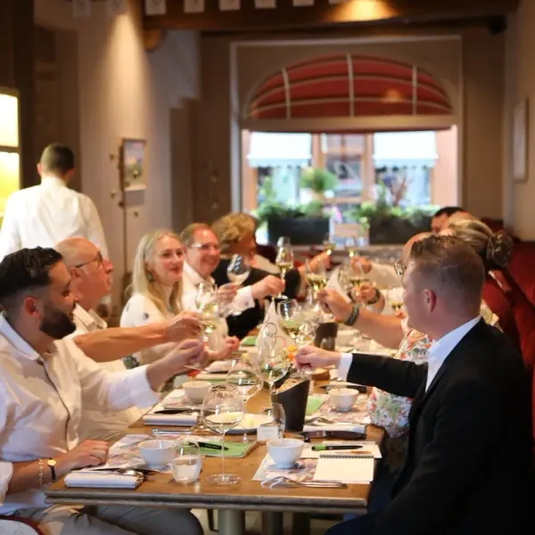 A group of people sitting around a table in a restaurant, sharing a meal and conversations, with glasses of wine and small bowls in front of them.