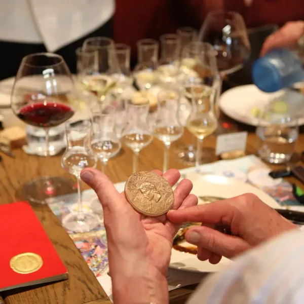 A person holds a gold medal in front of a table with wine glasses and documents.