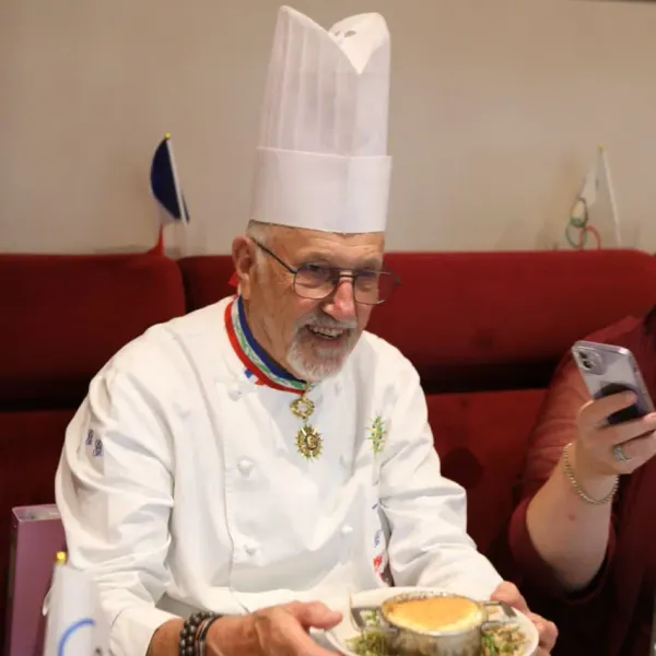 A chef in a white uniform with a tall hat and medals presents a dish to a person holding a smartphone.