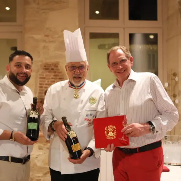 Three men pose together in a room, one in a chef's outfit with a medal, holding wine bottles, another with a red book.