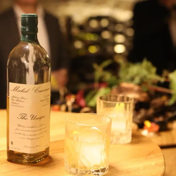 A bottle of Michel Couvreur "The Unique" whisky placed on a wooden table with two whisky glasses
