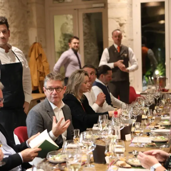 A group of people sitting around a table with glasses and dishes, a chef in a black apron standing to the left.