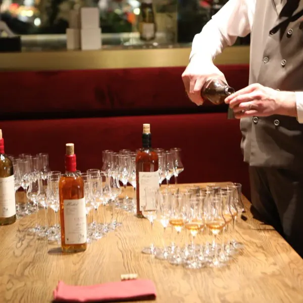 A server in a jacket pours wine into glasses lined up on a wooden counter, with several wine bottles nearby.