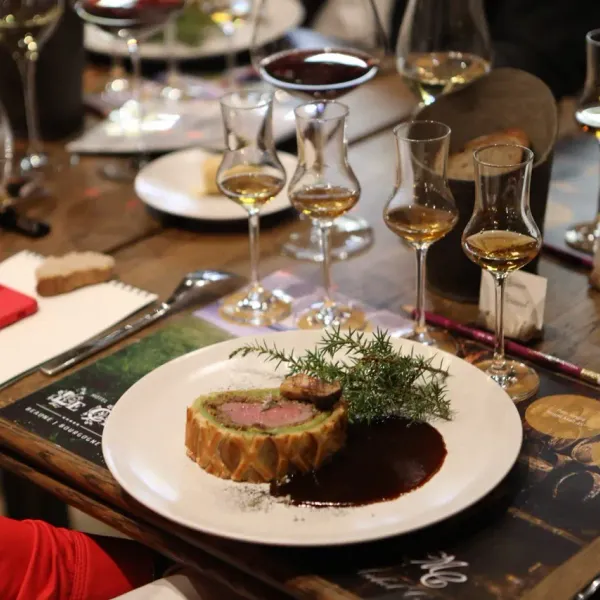 A meat pie with sauce and herbs, surrounded by wine glasses and utensils on a wooden table.