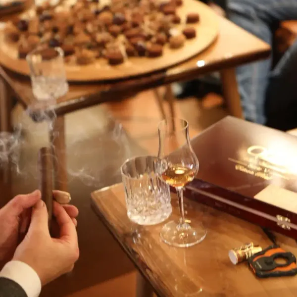 A person holds a lit cigar near a table with wine glasses, chocolates, and a bottle.
