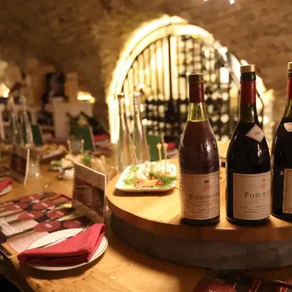 Bottles of Pomard wine on a wooden table in a lit wine cellar, accompanied by dishes and cutlery.