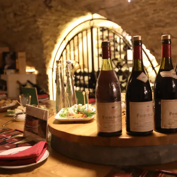 Bottles of wine and a plate of food on a wooden table in a lit wine cellar
