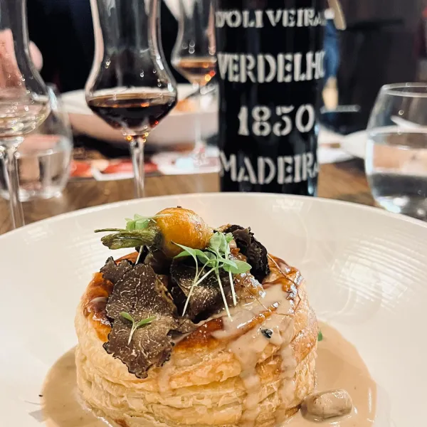 A gourmet dish featuring a thin tart topped with truffles and an egg yolk, served with two glasses of wine and a bottle of Madeira wine.