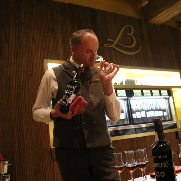 A man in a suit pours wine into a glass in an indoor setting with bottles and glasses on a wooden table.