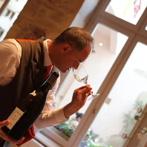 A man in a jacket and vest examines a glass of wine, holding a bottle in his hand, near an open window.