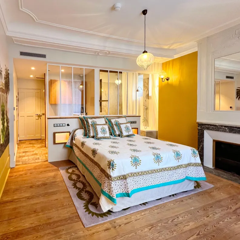 Chambre avec lit à baldaquin décoré de motifs floraux, cheminée en marbre, miroir et porte vitrée menant à une autre pièce