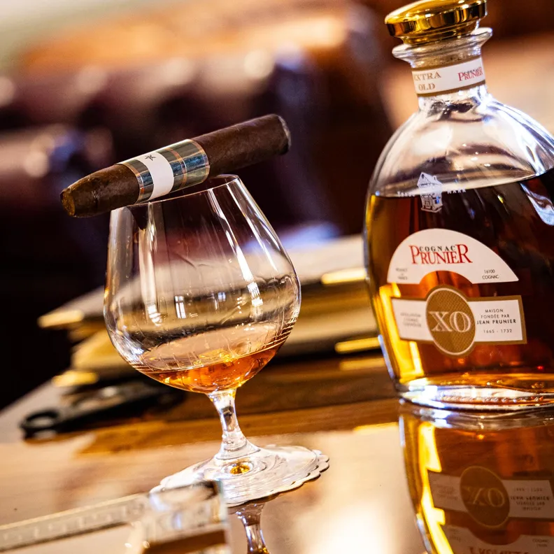 Une bouteille de cognac Premier avec un verre à dégustation contenant un liquide ambré et un cigare posé dessus sur une table en bois