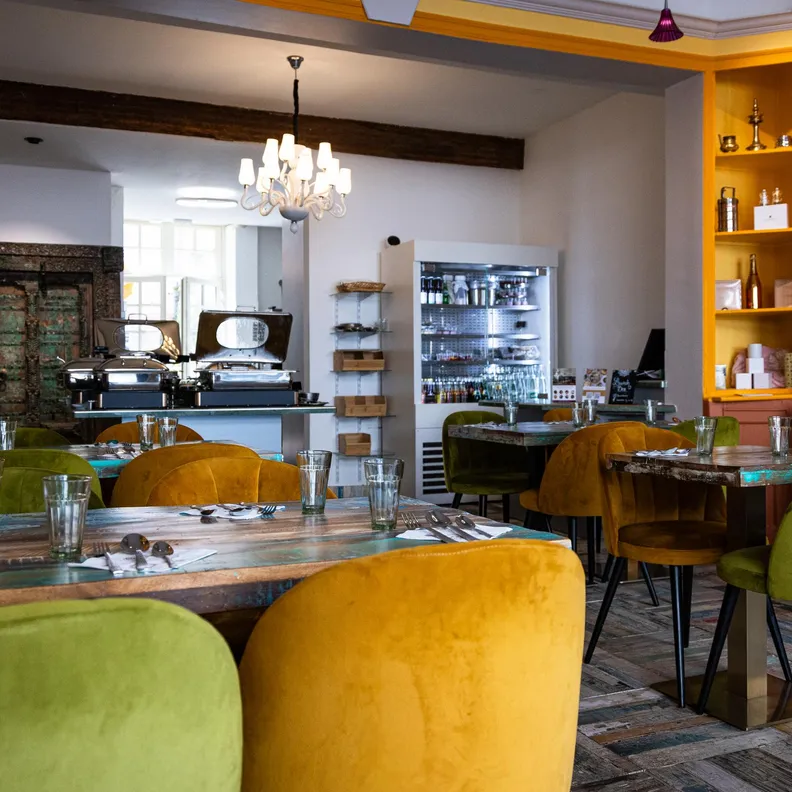 Restaurant intérieur avec tables et chaises colorées, comptoir et étagères remplies de bouteilles et d'objets décoratifs
