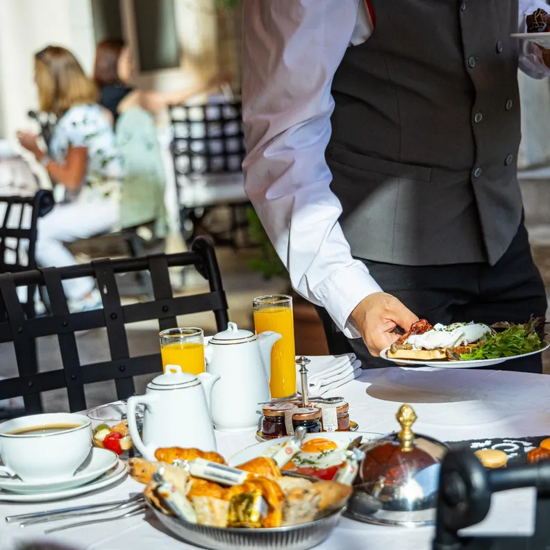 Un serveur en veste blanche et gilet gris pose une assiette de nourriture sur une table garnie de plats et de boissons