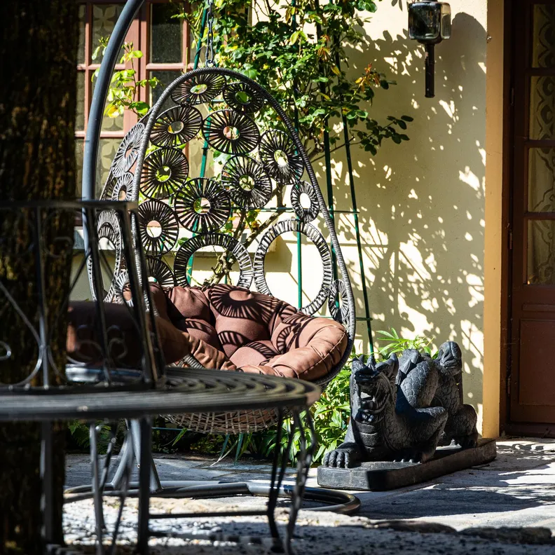 Un hamac décoratif avec des coussins est suspendu devant une porte en bois à panneaux de verre, entouré de plantes en pot et d'une table en fer forgé