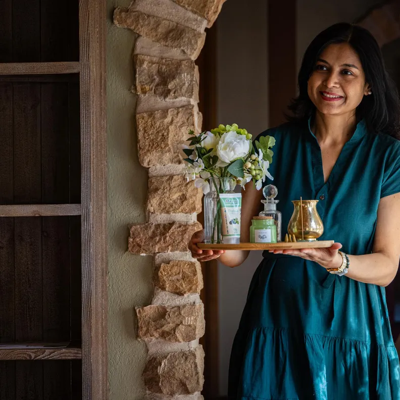 Une femme en robe verte tient un plateau avec une fleuriste et des produits de toilette près d'une fenêtre aux rideaux blancs