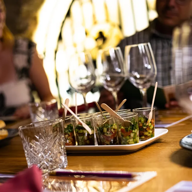 Une table avec des verres à vin, des petits plats contenant des amuse-bouches et des personnes en arrière-plan dans un cadre éclairé