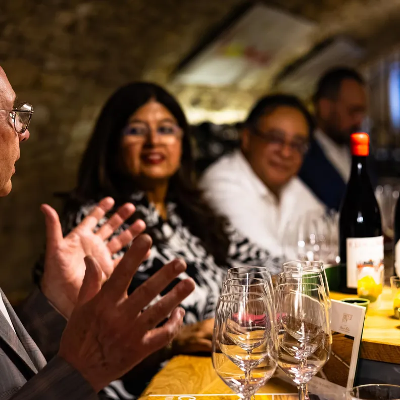 A man in a suit speaks animatedly to a group of people around a table with wine bottles and glasses.