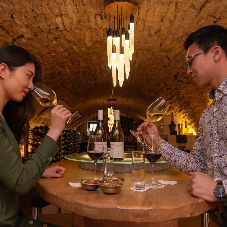 Two people sitting at a table are tasting wine in a wine cellar lit with bottles on the table and stone walls.