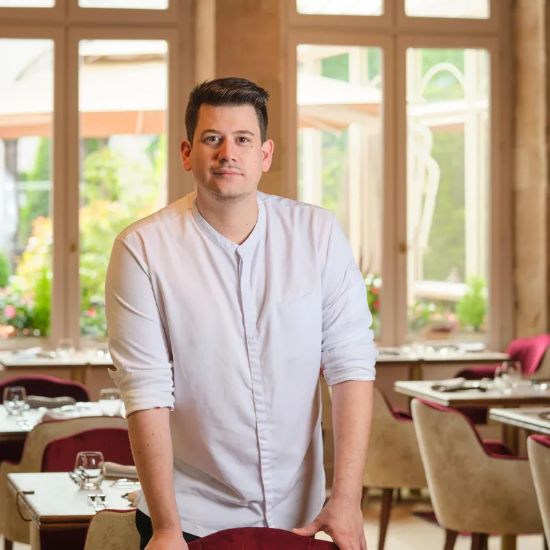 A man in a white shirt stands in a restaurant with set tables and large wooden windows.