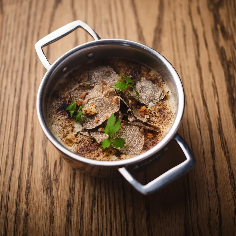 A metal pot containing soup with slices of truffle and herbs, placed on a wooden table