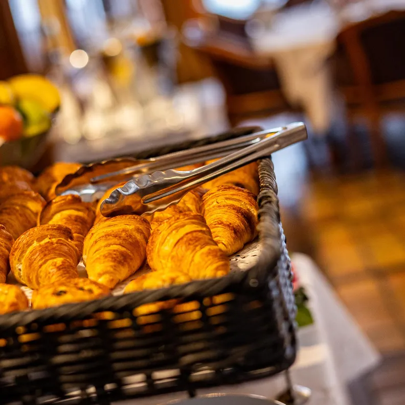 Un panier en métal contenant des croissants dorés avec des tongs pour les servir dans une boulangerie