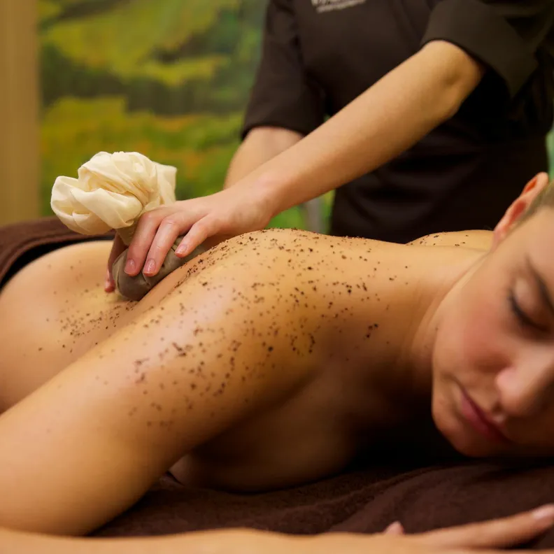 Une personne reçoit un massage à l'aide d'un gant de crins et d'un tissu enroulé, allongée sur le ventre avec la peau recouverte de particules noires