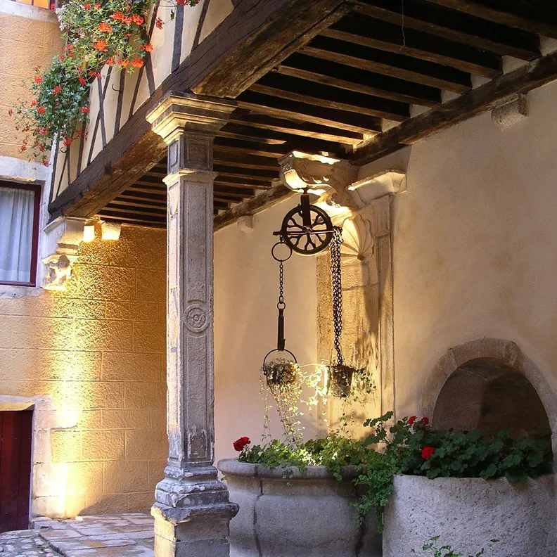 Un balcon en bois avec des colonnes en pierre, des plantes en pot et un mécanisme de poulie suspendu, éclairé par des lumières murales