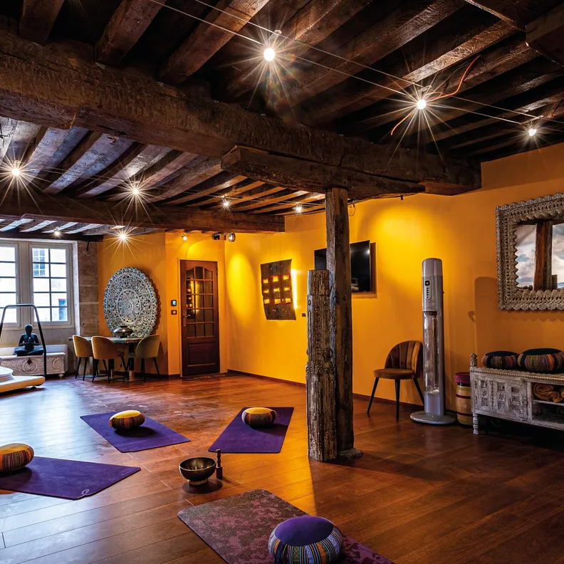 Salle de yoga avec des tapis au sol, des coussins, des murs jaunes et des poutres apparentes, éclairée par des spots au plafond
