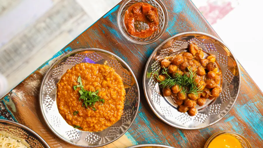 Three metal plates containing Indian dishes, placed on a wooden tablecloth with painted patterns, accompanied by rice and a glass of yellow liquid.