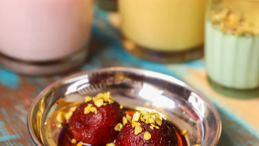 Two dark red dessert balls topped with pistachios in a small metal bowl, surrounded by glasses containing colorful drinks.