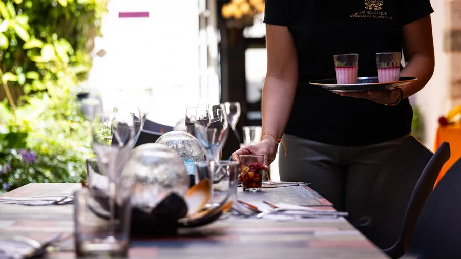 Une personne en noir sert des boissons roses sur un plateau dans un cadre extérieur avec des tables dressées