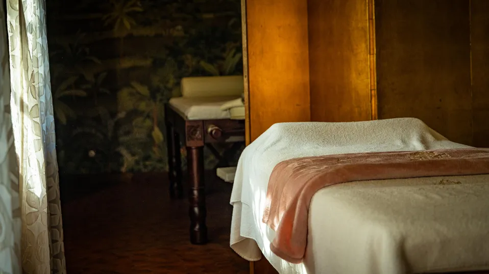 Un lit recouvert d'une couverture blanche et d'un plaid marron dans une pièce faiblement éclairée avec une table de chevet en bois