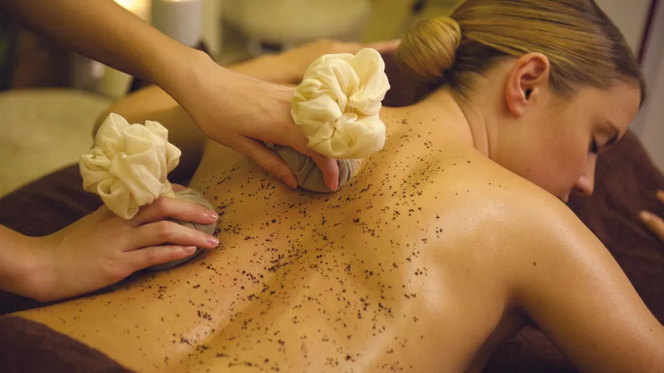 Une personne reçoit un massage du dos avec des fleurs et une poudre noire dans un cadre spa éclairé aux bougies